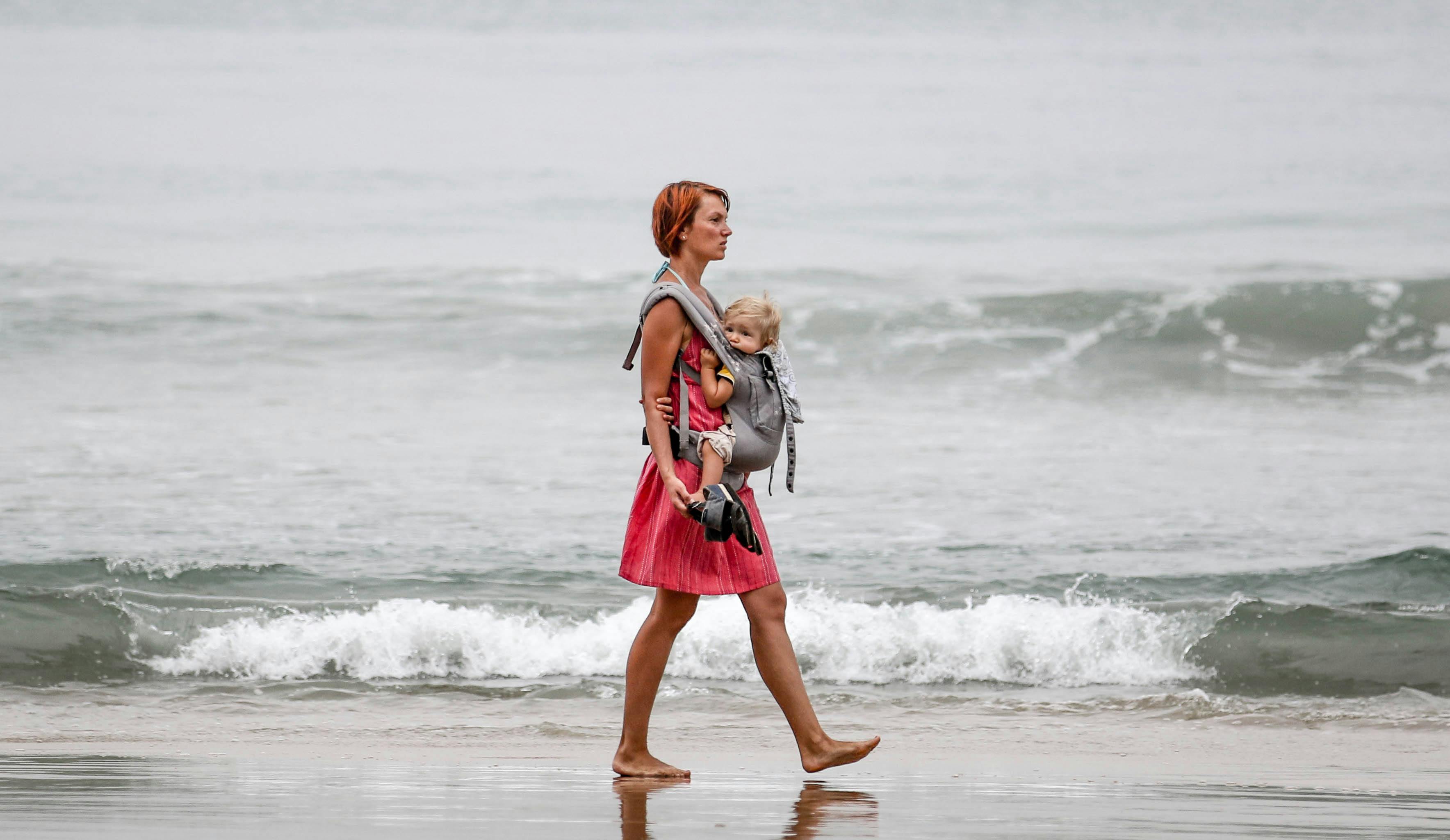 A woman carries her baby in a carrier while strolling along the serene Varkala Beach.