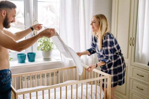 Free A smiling couple sets up a crib in a sun-filled room, preparing for a new baby's arrival. Stock Photo
