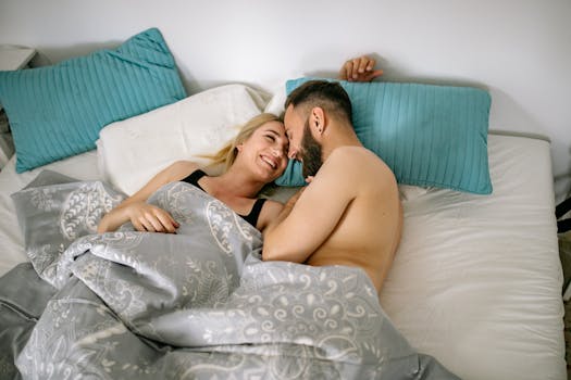 Smiling couple cuddling in bed under covers, enjoying intimate pillow talk.