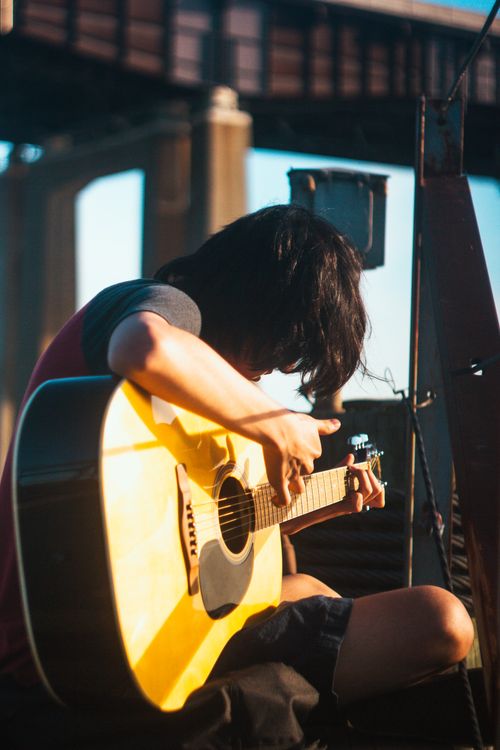Free Young adult with black hair playing an acoustic guitar outdoors under a bridge. Stock Photo
