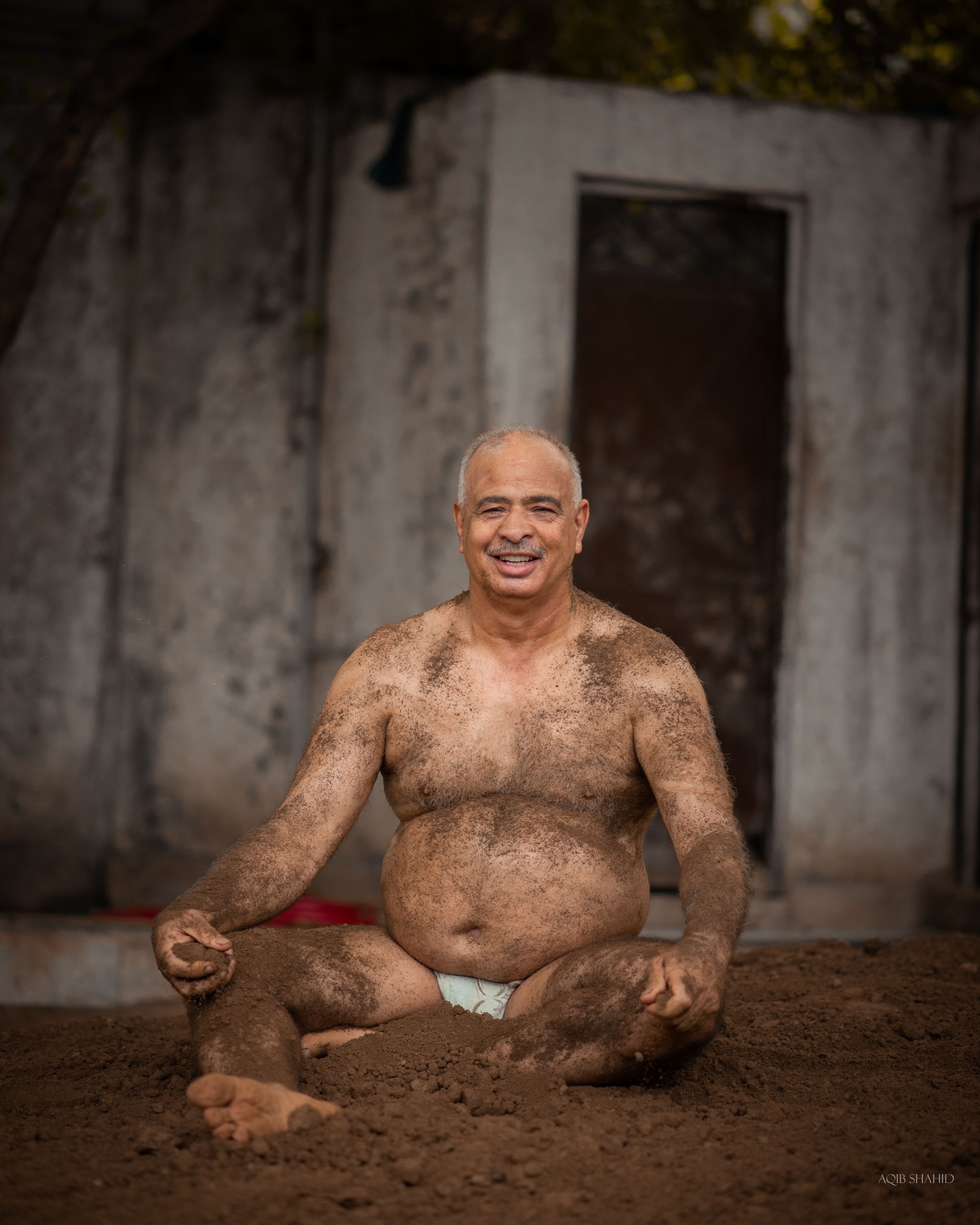 An adult man covered in mud, sitting outdoors, embracing a traditional wrestling ritual.