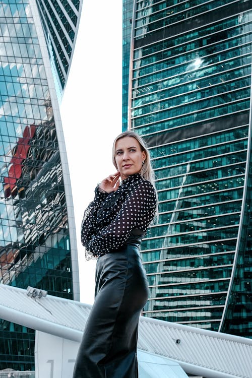 Side view confident female in posh outfit standing against contemporary skyscrapers while touching chin and looking away thoughtfully