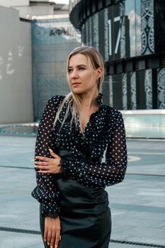 Confident young female in classy outfit touching arm and standing outside contemporary downtown building while looking away