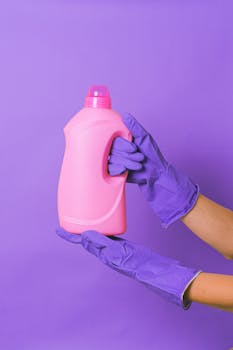 Pink detergent bottle held by gloved hands on purple background, studio shoot.