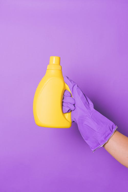 Unrecognizable person in protective glove with plastic bottle of chemical cleaning fro daily routine in hand on purple background in studio