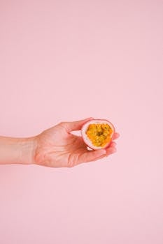 A hand holding a half passion fruit against a vivid pink background, showcasing its fresh and exotic appeal.