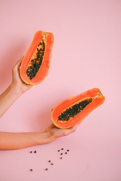 Crop unrecognizable female showing halves of tasty exotic papaya fruit against pink background above fallen papaya seeds