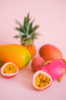 Colorful tropical fruits including pineapples, mangoes, and dragon fruit displayed on a pastel pink surface.