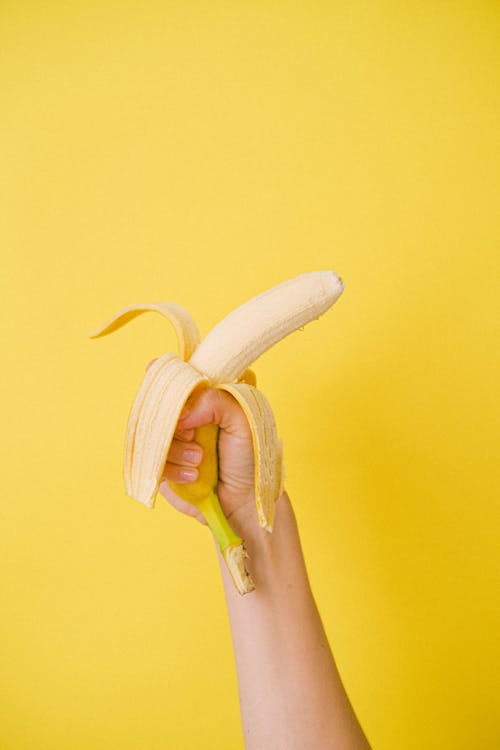 Crop anonymous female holding half unpeeled ripe organic banana against bright yellow background before eating