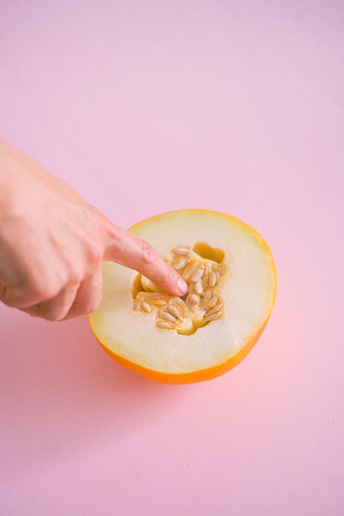 From above crop anonymous person touching gently ripe delicious melon cut into halves placed on pink surface