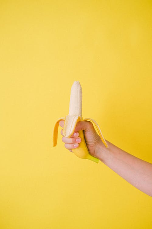 Crop anonymous female showing sweet half unpeeled banana against bright yellow wall in studio