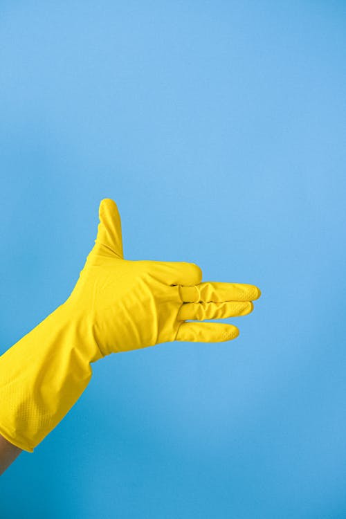 Crop anonymous person in yellow latex glove demonstrating finger gun gesture against blue wall