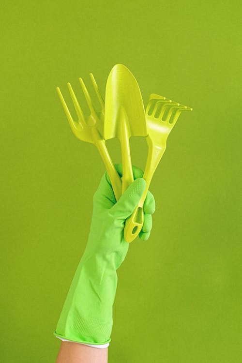 Crop anonymous person wearing rubber glove showing set of gardening tools including trowel and hand rake against green background