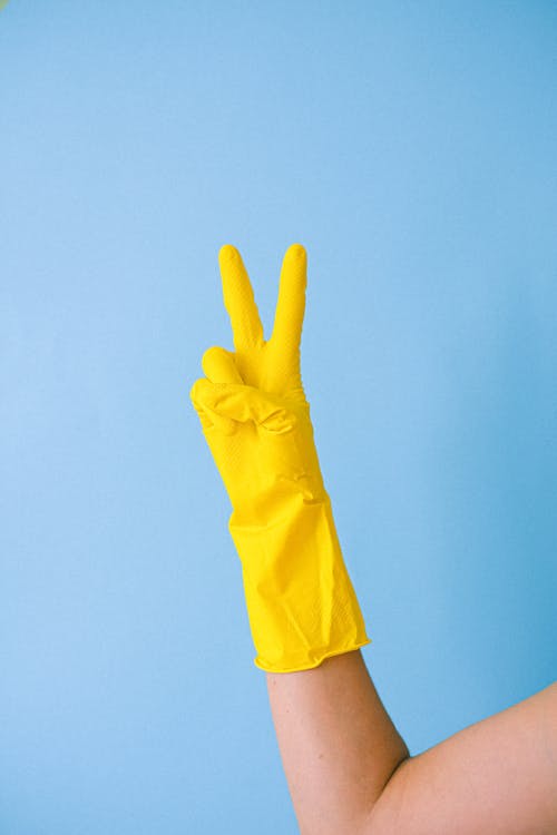 Crop hand of anonymous person in rubber gloves for cleaning demonstrating peace symbol against blue background