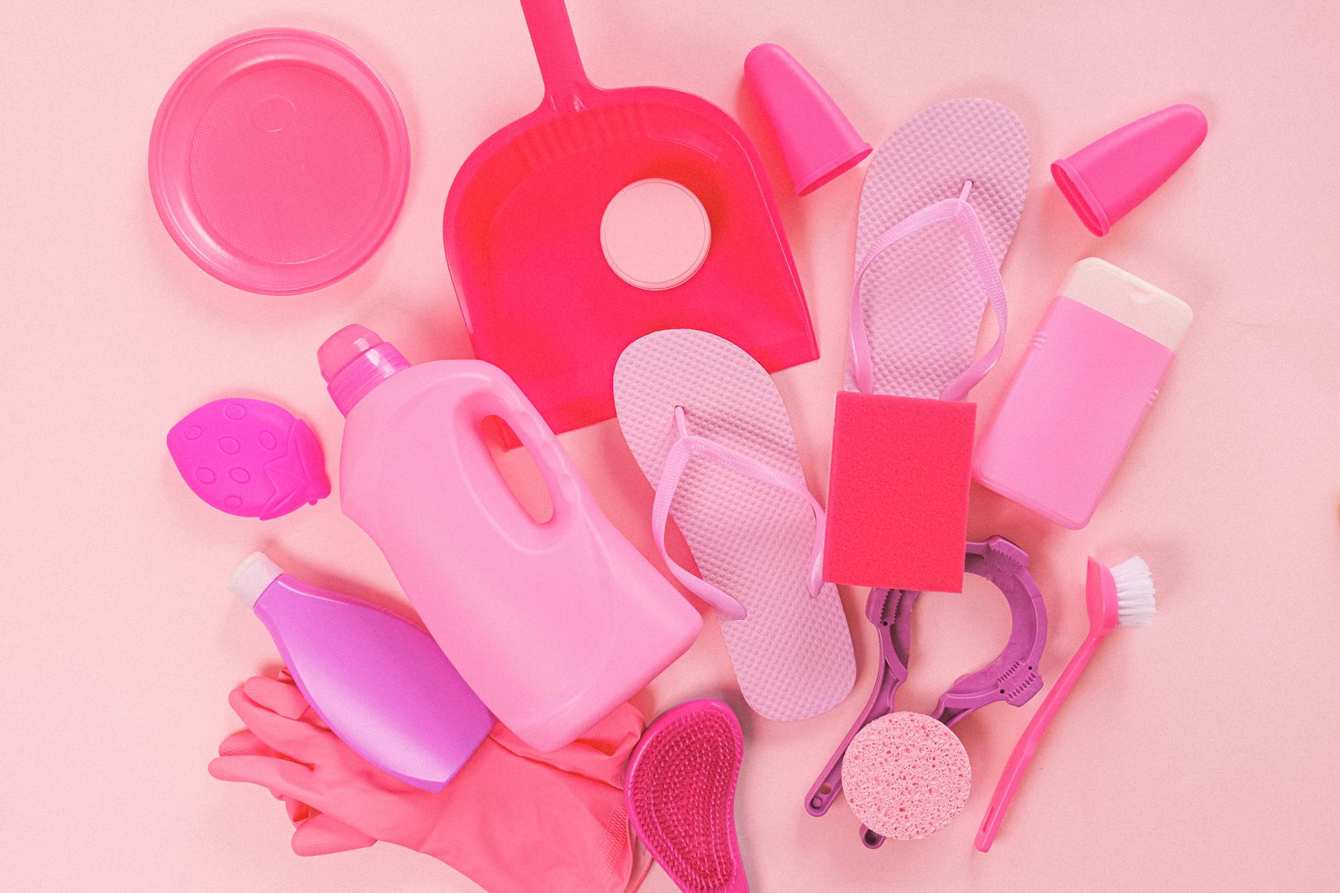 Collection of various plastic detergent bottles and cleaning tools placed on pink background near flip flops and rubber gloves
