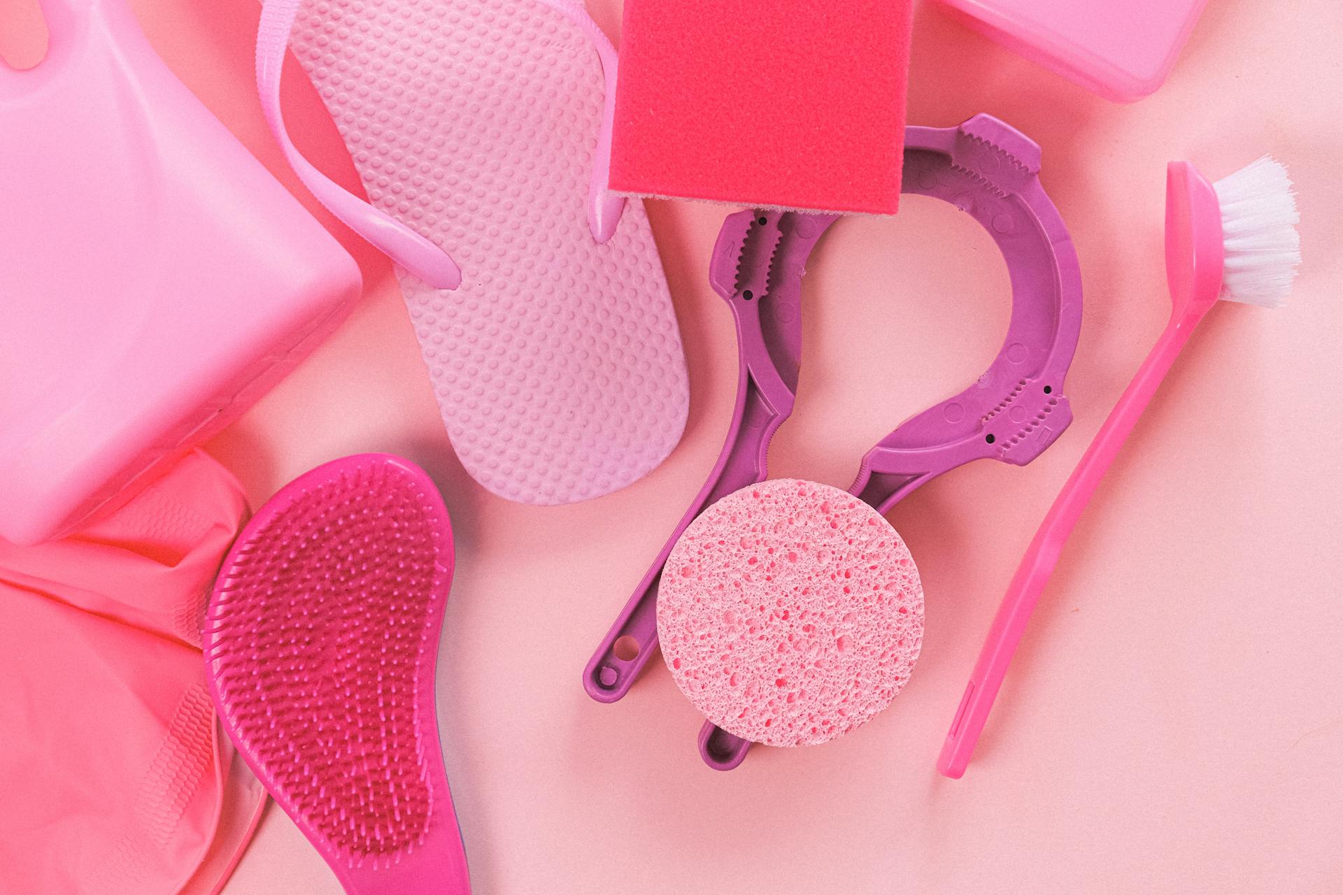 Overhead layout of assorted cleansing tools including brush sponge and plastic bottle arranged on light pink surface