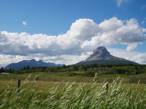 Scenic view of a mountain range with lush fields and a fence under a cloudy sky.
