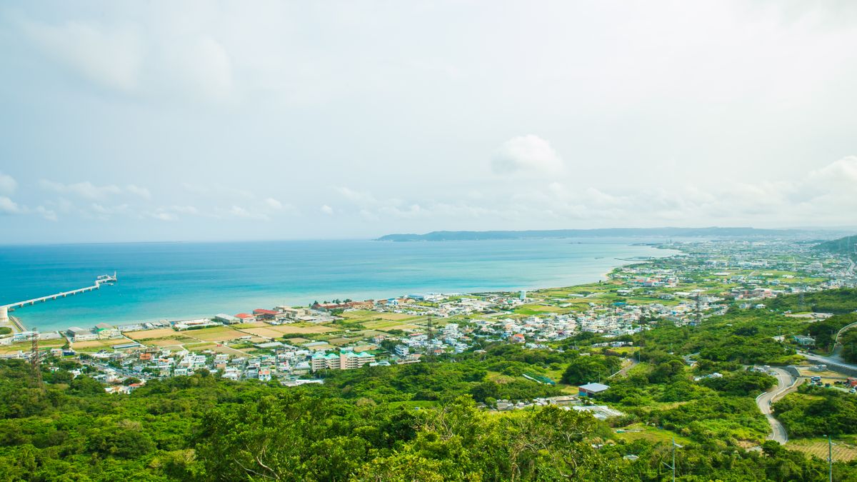 오키나와 — Calm turquoise water of ocean washing shore with small town and green tropical forest under cloudy sky