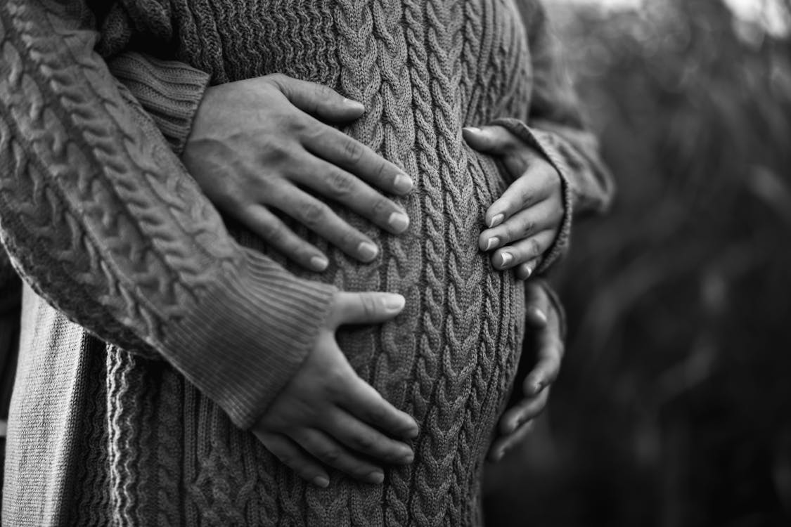 Black and white of crop anonymous male embracing belly of expectant girlfriend in knitted wear in park