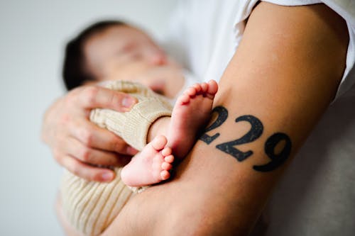 Side view of crop anonymous parent with tattoo holding adorable sleeping barefooted baby in arms at home