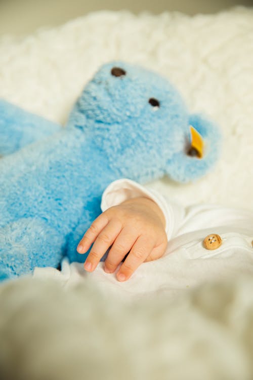 From above of crop unrecognizable baby in white pajama lying on soft bed with blue plush toy in daylight