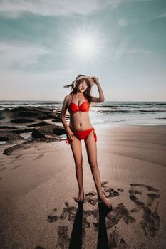 Stylish woman in red bikini enjoying a sunny day on the beach with hat and ocean backdrop.