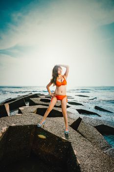 Woman in orange bikini standing on beach rocks with ocean background and bright sky overhead.