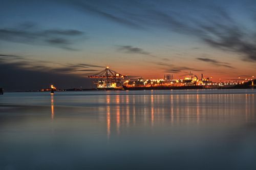 Free A scenic view of Gothenburg harbour at dusk with glowing cranes and calm sea reflections. Stock Photo