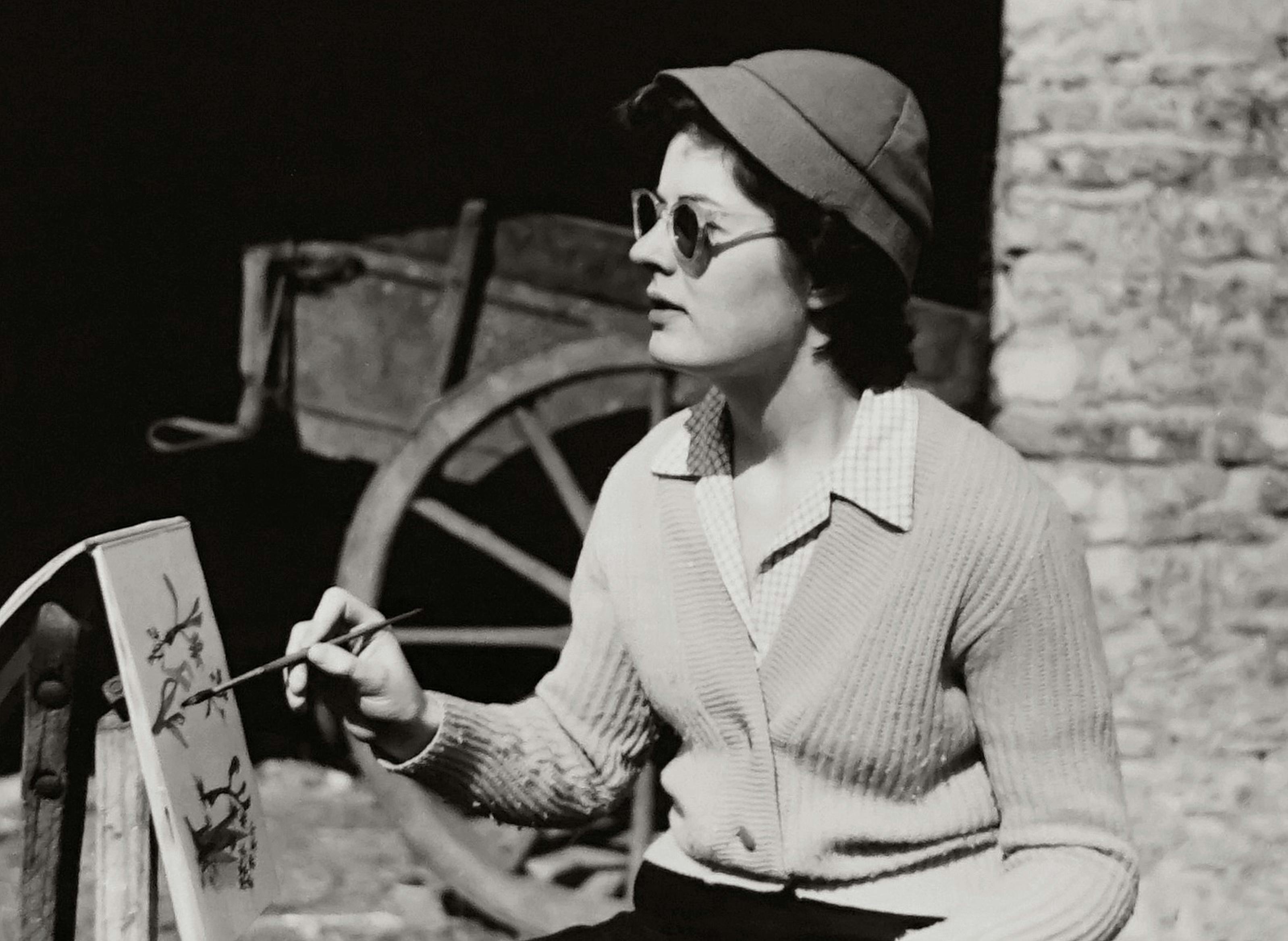 A retro monochrome portrait of a woman painting outdoors wearing sunglasses and a knitted sweater.