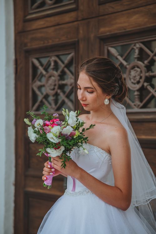Free Young bride in white dress standing near massive wooden door and holding bouquet of flowers at daylight Stock Photo