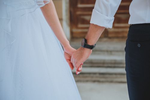Free Back view of unrecognizable arms of newlywed couple in wedding clothes holding hands while standing on street with blurred stair on background during celebration Stock Photo