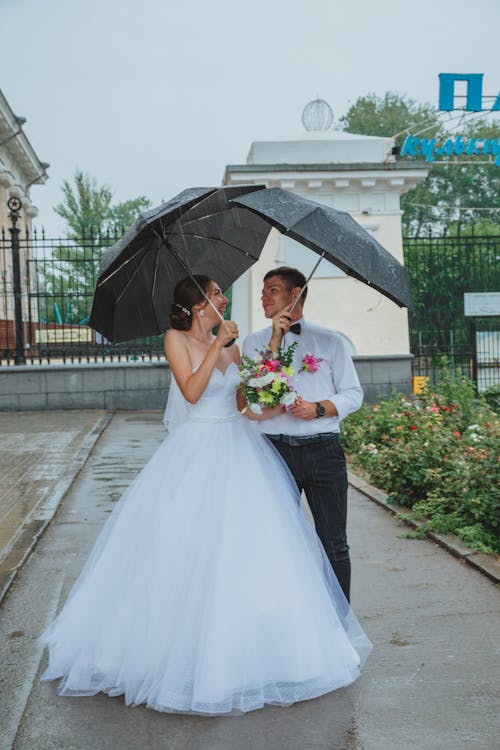 Full body of happy groom and bride in wedding outfit with bouquet of flowers looking at each other while standing on pavement under umbrellas in rainy weather