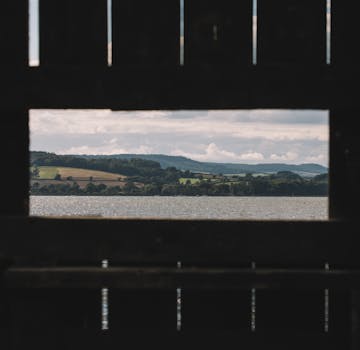 A serene landscape of a lake surrounded by rolling hills viewed through a wooden fence.