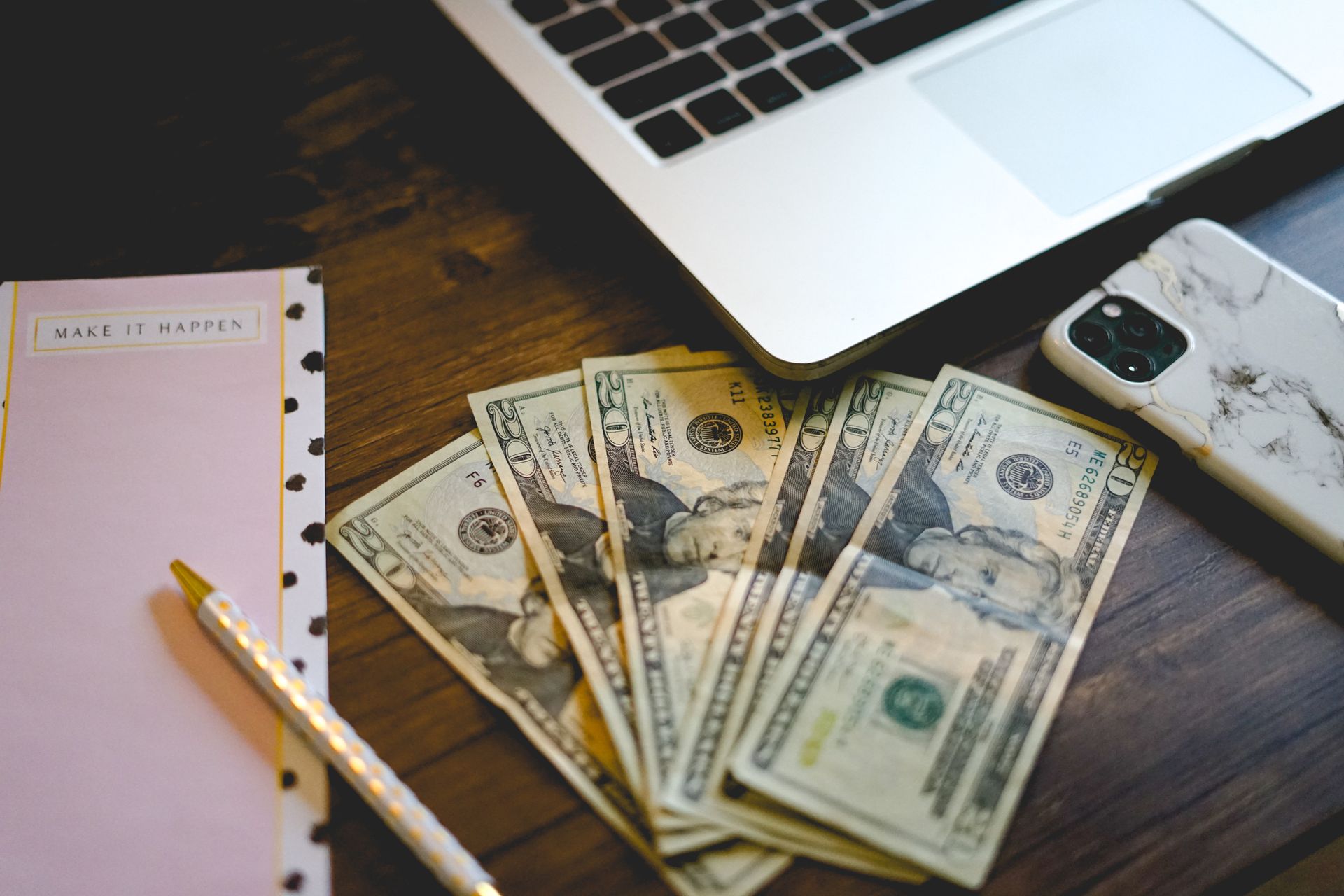 A workspace setup with dollar bills, notepad, and laptop on a wooden desk.