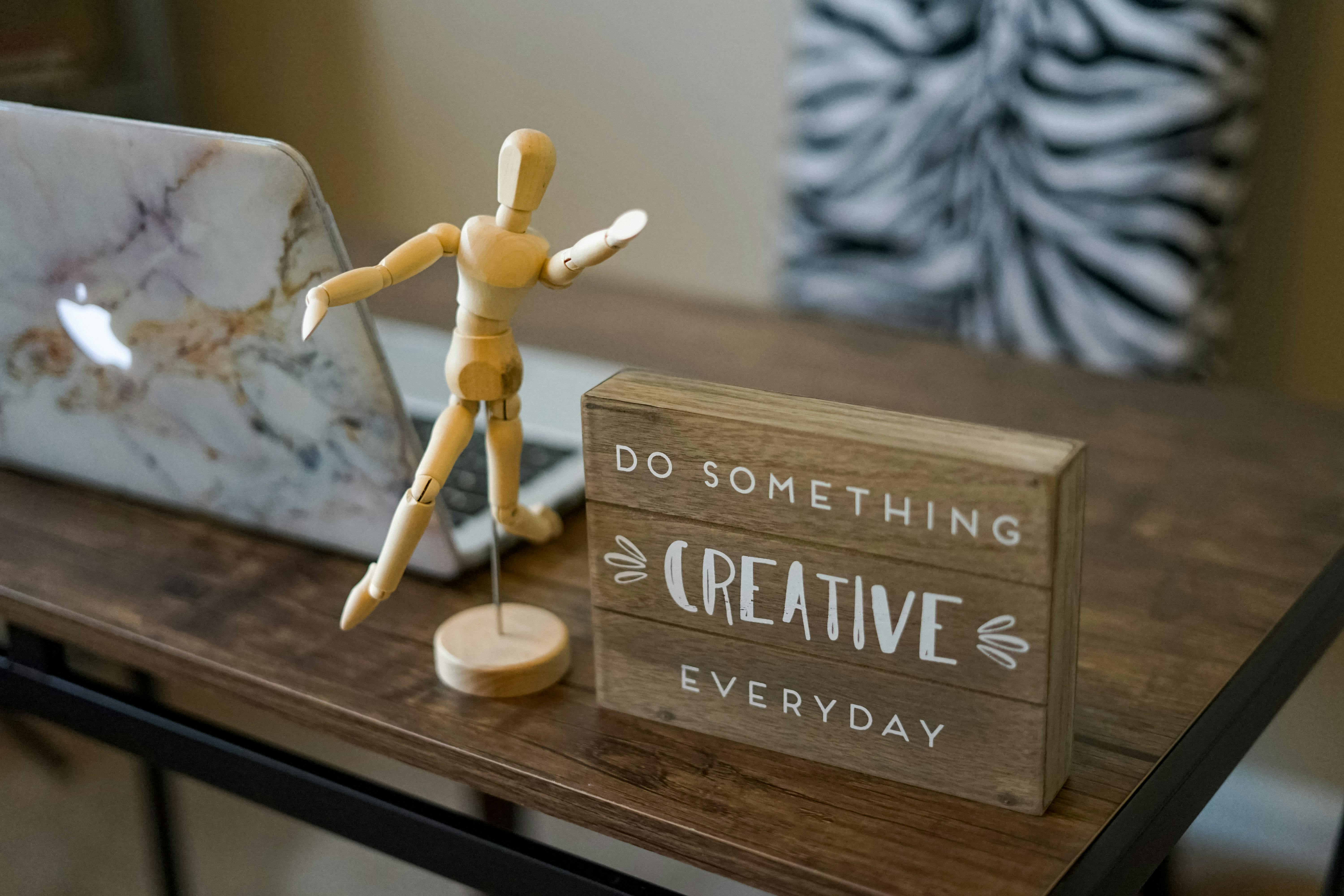 Wooden mannequin and inspirational sign on a desk beside a laptop, promoting creativity.