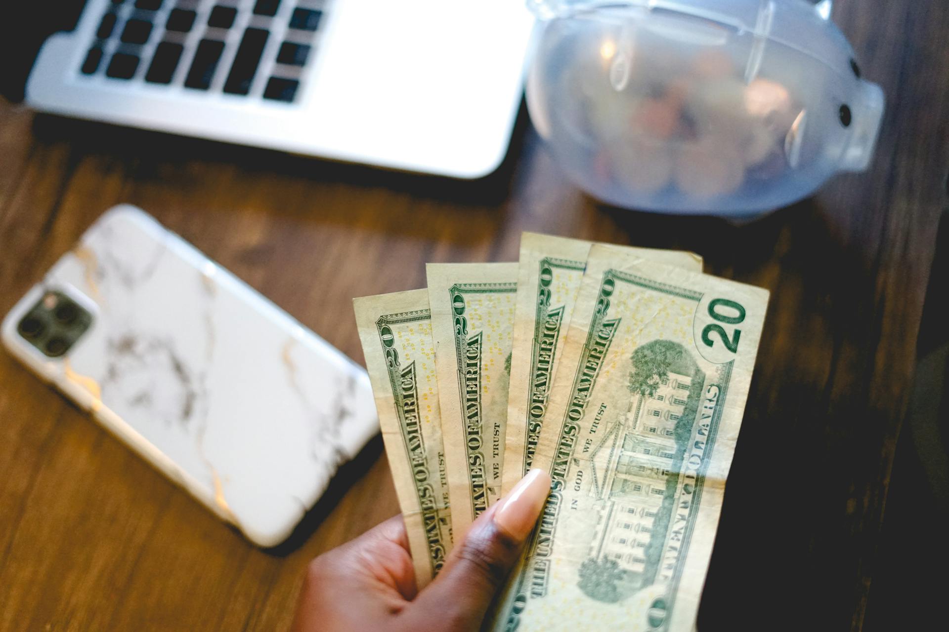 Close-up of a hand holding dollar bills beside a laptop and piggy bank, symbolizing savings and finance.