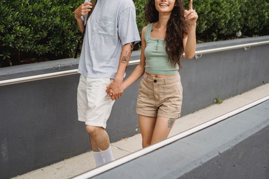 Crop unrecognizable couple holding hands and walking on city street against green plants in daytime
