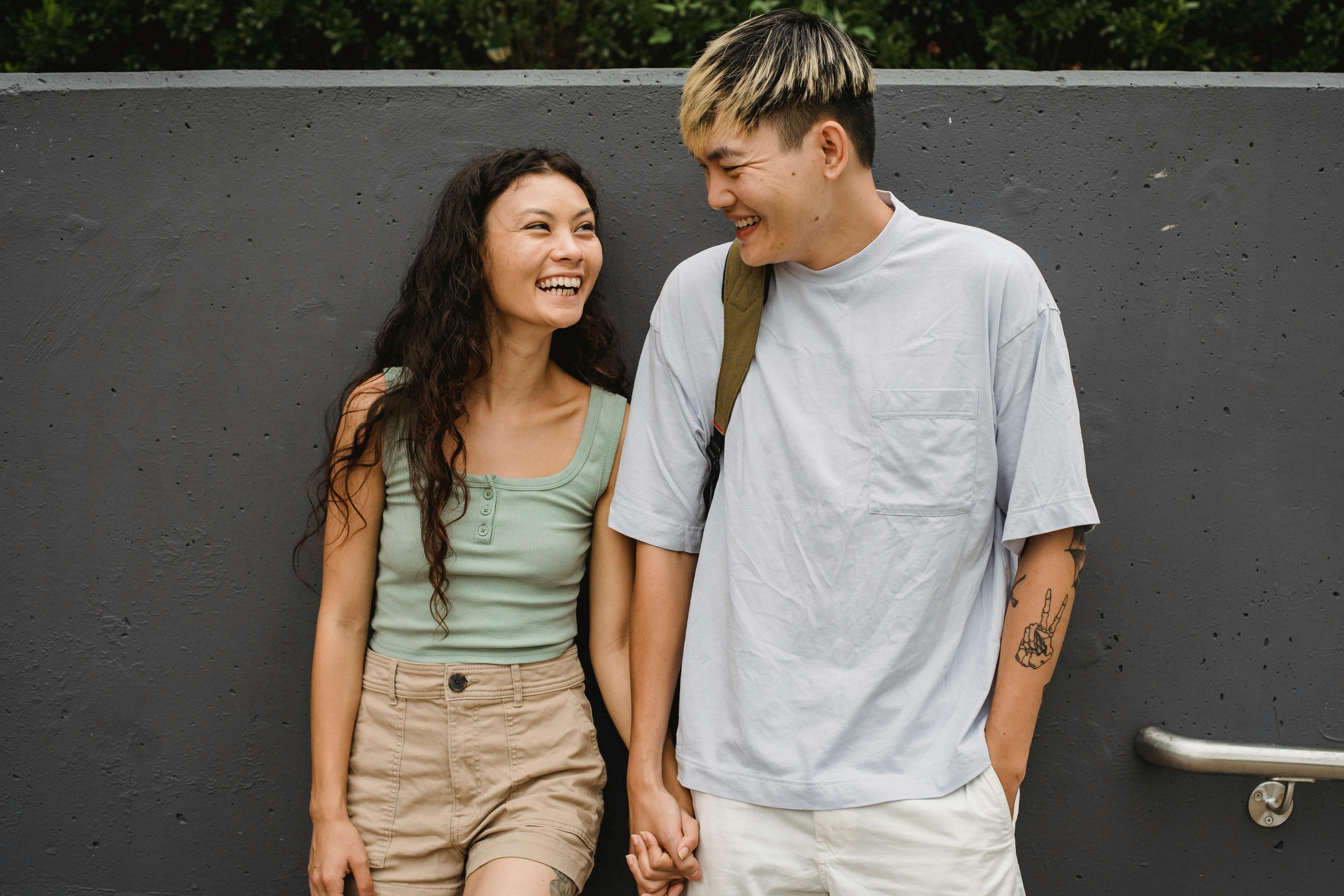 Delighted diverse couple standing together and looking at each other against gray stone wall in daytime