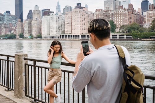 Young man photographing girlfriend on smartphone during date in city downtown near river