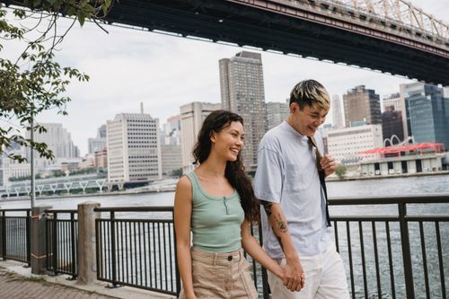 Free Positive young Asian man and ethnic woman holding hands and smiling while walking together on quay near bridge over river in New York Stock Photo