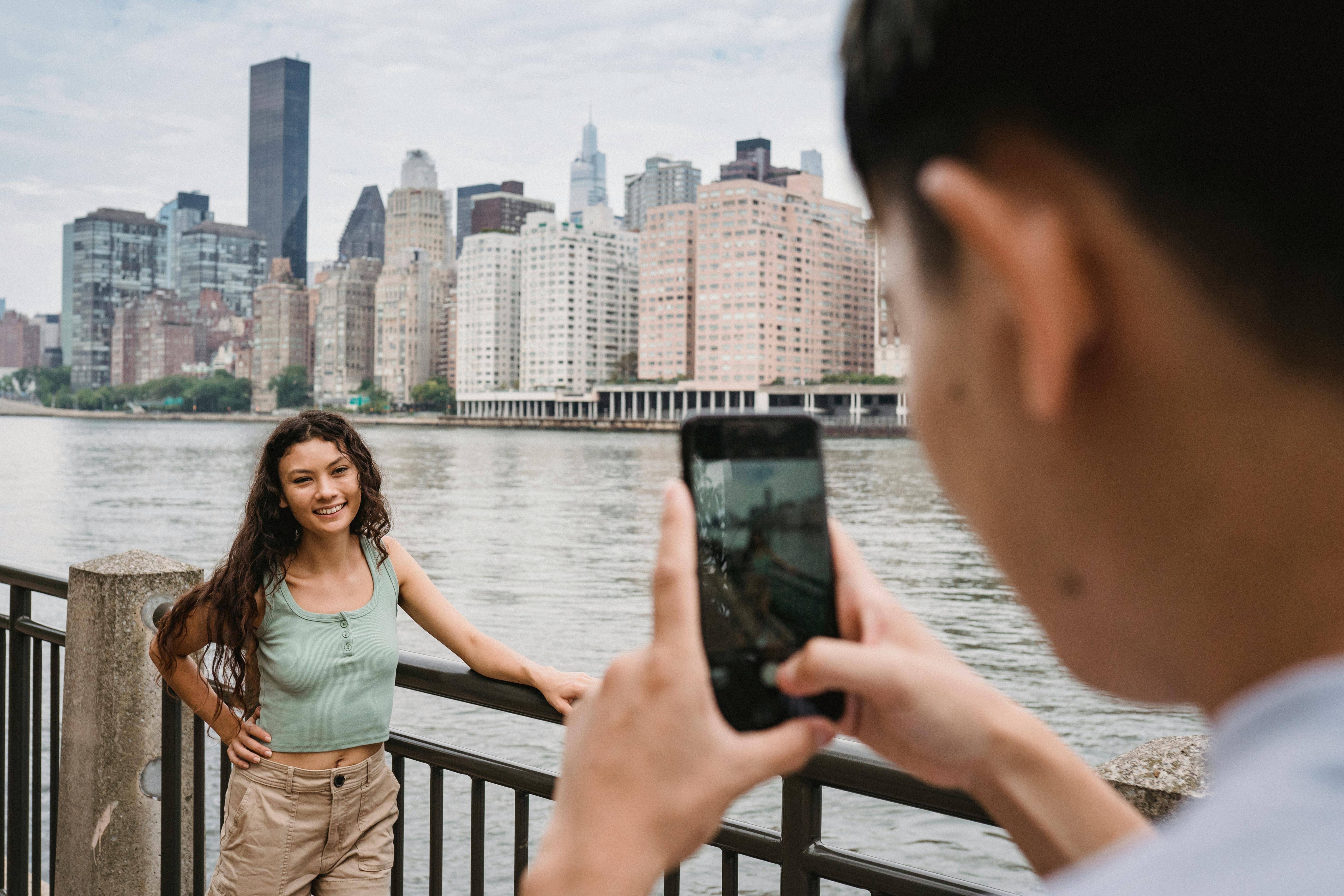 crop male taking photo of smiling ethnic girlfriend on city embankment against modern skyscrapers