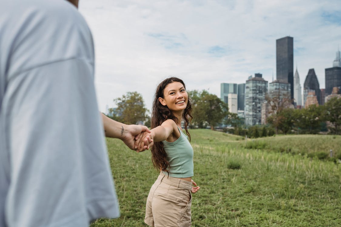 Free Crop anonymous man holding hand and following happy young ethnic girlfriend while spending time together in green park in New York Stock Photo