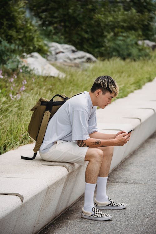 Chico étnico Joven De Moda Descansando En Un Banco En El Parque Y Con Smartphone