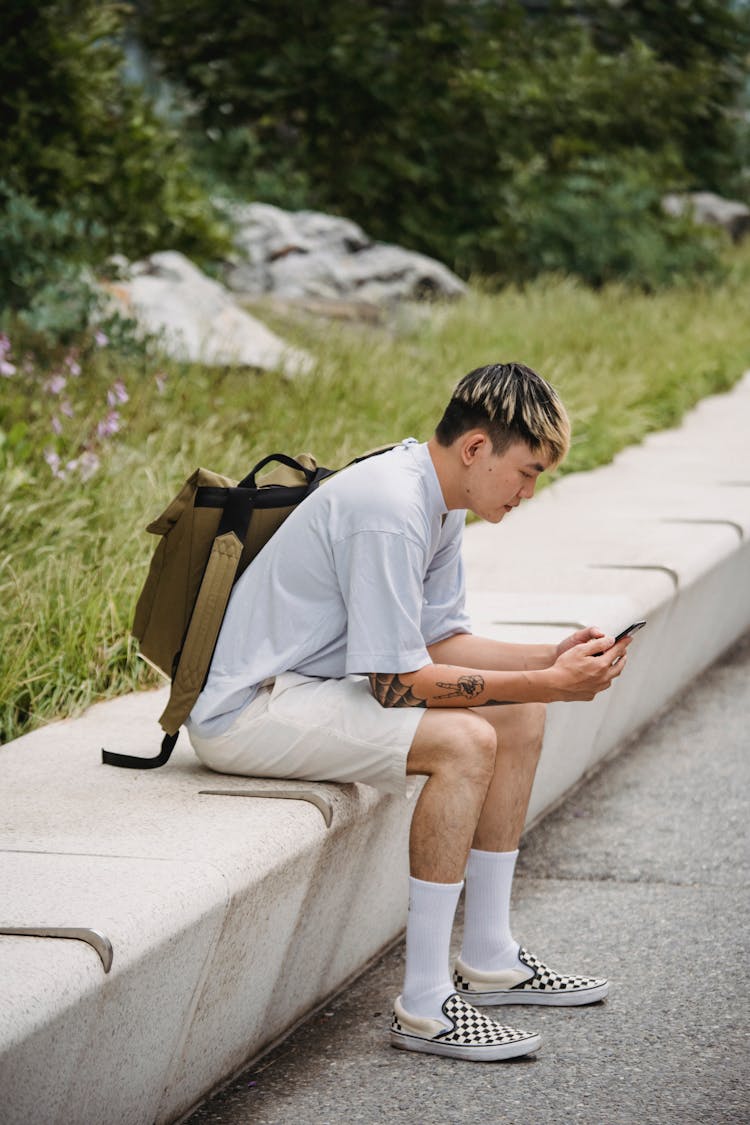 Trendy Young Ethnic Guy Resting On Bench In Park And Using Smartphone