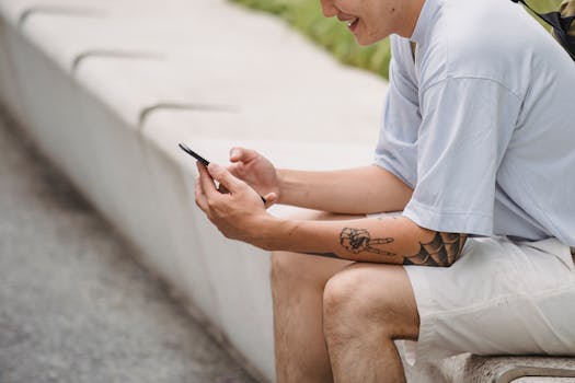 一名年轻的匿名纹身男性，穿着休闲服，面带微笑地坐在公园长凳上，用智能手机观看有趣的视频