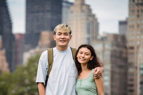 Cheerful Asian man with dyed hair embracing smiling ethnic girlfriend while looking at camera in town