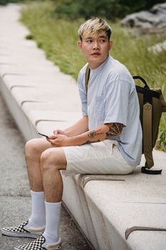 一位穿着休闲装、背着背包、拿着手机的年轻少数族裔男性坐在长凳上，目光移向别处，思考着纹身的事
