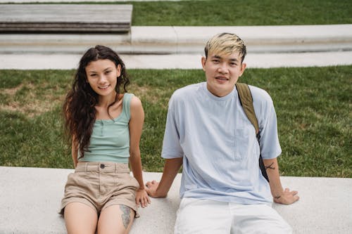 Content multiracial couple in casual wear with tattoos sitting on bench while looking at camera in daylight