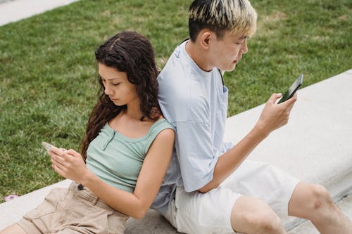 Free From above of crop diverse couple text messaging on cellphones while sitting on bench near lawn in daylight Stock Photo