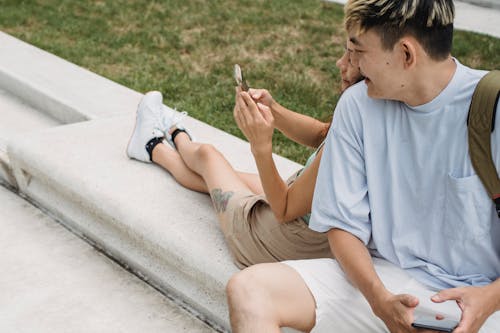 From above of tattooed ethnic female with crossed legs showing cellphone to crop cheerful beloved while sitting on bench in city